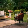 A woman sits in a hanging chair in a garden, holding a mug and looking at her phone. A small table with breakfast items and flowers is nearby. Voir les images du produit