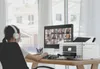 A person wearing headphones is working on a computer in a home office setting, with a printer next to the monitor. Náhled obrázku produktu