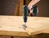 A hand is using a cordless drill to screw a metal bracket onto a wooden workbench. Produktbild-Vorschau