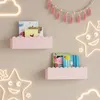 Two pink scalloped shelves mounted on a wall, holding children's books and decorative items. Neon cloud and star lights are also visible. Produktbild-Vorschau