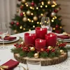 A Christmas-themed table centerpiece with red LED candles, pine branches, berries, and gold ornaments, set on a wooden slice, with a blurred Christmas tree in the background. Produktbild-Vorschau