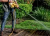 A person is using a pressure washer on a wooden deck next to a lawn and bushes. Podgląd zdjęcia produktu