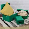 Three green storage boxes of different sizes are arranged on a rug, with a yellow cushion and stuffed toys inside two of them. The background is a brick wall. Produktbild-Vorschau