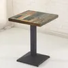 A square table with a distressed wood-look top and a dark metal pedestal base, set against a white brick wall and light-colored floor. Produktbild-Vorschau