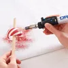 A person is using a Dremel VersaTip torch to heat a red and white striped object held on a wooden stick, causing red powder to scatter. Produktbild-Vorschau