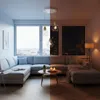 A split image showing a modern living room during the day and night, featuring a sectional sofa, coffee table with candles, and a pendant light fixture. Produktbild-Vorschau