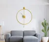 A modern gold wall clock with a pendulum hangs on a white wall above a grey sofa. A cactus plant is visible to the right. Náhľad obrázku produktu