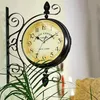 A vintage-style double-sided wall clock mounted on a wall, with a plant in the foreground. Produktbild-Vorschau