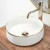 A white ceramic basin with a gold rim sits on a wooden surface, with a gold faucet above it. Produktbild-Vorschau