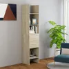 A tall, narrow bookshelf with two drawers and several shelves, placed against a light gray wall. The bookshelf is filled with books and decorative items. A large green plant is visible to the right, and a portion of a painting is on the wall to the left. Produktbild-Vorschau