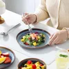 A person is eating a salad from a blue plate, with other fruit dishes and a glass of water on a white table. Náhled obrázku produktu