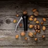 A black and silver nutcracker is placed on a rustic wooden surface, surrounded by various nuts like walnuts, almonds, and hazelnuts. Produktbild-Vorschau