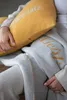 A person is holding a yellow pillow with text and is covered by a gray blanket with text. Produktbild-Vorschau