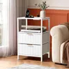 A white nightstand with two drawers and an open shelf is placed next to a sofa in a living room. The nightstand has a turntable on the shelf and electronic devices on top. The sofa is covered with a brown throw blanket. Náhled obrázku produktu