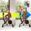 A woman is exercising on a stationary bike in a bright room with a large window overlooking trees. The image is split into two halves, showing the woman in different positions on the bike. An inset diagram highlights adjustable handlebars. Produktbild-Vorschau