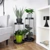 Two black metal plant stands with plants on a wooden floor next to a gray armchair and a white bookshelf. Produktbild-Vorschau