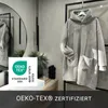 A fluffy grey hoodie blanket is displayed in a modern bathroom setting, with a mirror and sink visible. Produktbild-Vorschau