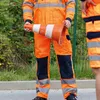 Two people in orange work clothes stand outdoors, one holding a traffic cone. Anteprima dell’immagine prodotto