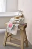 A stack of floral patterned towels and a brush are placed on a wooden stool next to a bathtub. Produktbild-Vorschau