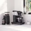 Two black coffee machines and a coffee grinder are displayed on a white countertop, with a window in the background. Produktbild-Vorschau