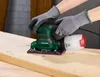 Hands wearing gray gloves are holding a green and white orbital sander, sanding a wooden plank. Produktbild-Vorschau