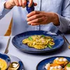A person is seasoning a plate of ravioli with a pepper grinder. Other plates of food are visible. Voir les images du produit
