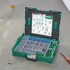 A green toolbox filled with various screws, bolts, and wall plugs, resting on a concrete floor. Podgląd zdjęcia produktu