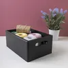 A black wooden crate filled with spools of twine and other craft supplies, placed on a white surface with a maroon wall in the background and a potted plant to the right. Produktbild-Vorschau
