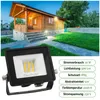 A black LED floodlight is shown in the foreground, with a wooden cabin and garden in the background. Produktbild-Vorschau