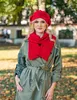A woman wearing a red beret and scarf stands in a park with autumn leaves on the ground. Anteprima dell’immagine prodotto