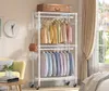 A white wire clothing rack with clothes and a suitcase, positioned in a room with a yellow chair, a clock, and a window. Voir les images du produit