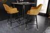 Two mustard yellow velvet bar stools with black legs and gold accents are placed around a dark wood bar table in a modern interior setting. Produktbild-Vorschau
