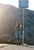 A woman stands by a signpost in a mountainous landscape. Produktbild-Vorschau