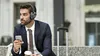 A man in a suit wearing headphones and holding a phone sits next to a suitcase. Produktbild-Vorschau
