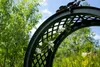 A garden arch with white flowers and green leaves against a backdrop of trees and a blue sky. Produktbild-Vorschau