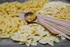 A variety of dry pasta shapes are arranged on a wooden surface, with a wooden spoon holding some fusilli pasta in the center. Produktbild-Vorschau