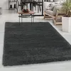 A dark gray shaggy rug is shown in a living room setting with a coffee table, chairs, and a sofa in the background. Produktbild-Vorschau