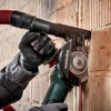 A person wearing gloves is using a Metabo angle grinder with a dust extraction hood attached to cut a groove into a brick wall. Produktbild-Vorschau