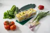 A glass baking dish filled with pasta and vegetables, surrounded by fresh ingredients like tomatoes, celery, parsley, cucumber, red bell pepper, and green onions. Produktbild-Vorschau