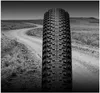 A close-up of a bicycle tire in the foreground, with a winding dirt road and grassy hills in the background. Produktbild-Vorschau