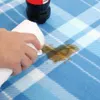 A hand is wiping a spill on a blue and white checkered picnic blanket with a white towel. A bottle of Coca-Cola is visible in the background. Produktbild-Vorschau