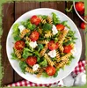A close-up overhead shot of a pasta salad on a white plate, placed on a wooden table with a red and white checkered cloth. Produktbild-Vorschau