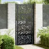 A black metal garden gate with a leaf design, set in a garden with brick pillars and greenery. Náhled obrázku produktu