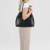 A woman is shown from the side, wearing a white t-shirt and beige pants, and carrying a black tote bag. Produktbild-Vorschau