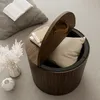A round wooden storage table with its lid open, revealing a pillow inside. Books are placed on top of the lid and next to a vase of dried flowers. Anteprima dell’immagine prodotto