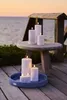 Several white LED candles are arranged on a wooden table and a blue tray, with a body of water and a wooden structure in the background. Produktbild-Vorschau