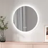 A round LED mirror with a light border is mounted on a wall above a light pink dresser. A vase with a green plant is to the left of the mirror, and a framed picture is to the right.