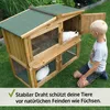 A boy is kneeling on the grass, looking at a wooden rabbit hutch with two rabbits inside. The hutch has a green roof and wire mesh windows. Produktbild-Vorschau