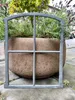 A gray metal window frame is placed in front of a large stone planter filled with green plants and lavender. Produktbild-Vorschau