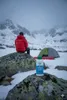 A person in a red jacket sits on a rock in a snowy mountain landscape, with a tent and a bottle of Nikwax Down Wash in the foreground. Náhled obrázku produktu
